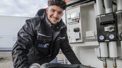 Ein junger Mann in Arbeitskleidung lächelt, während er an einer technischen Ausrüstung mit Druckmessgeräten arbeitet.