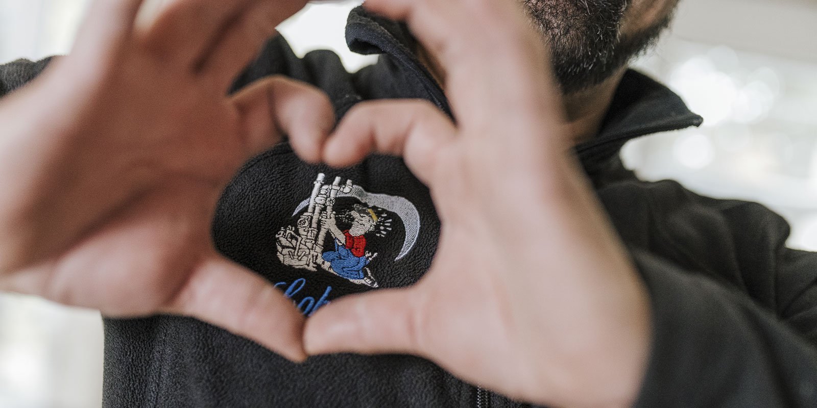 Eine Person formt mit ihren Händen ein Herz, während sie ein gesticktes Motiv auf ihrem schwarzen Pullover zeigt.