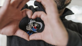 Eine Person formt mit ihren Händen ein Herz, während sie ein gesticktes Motiv auf ihrem schwarzen Pullover zeigt.
