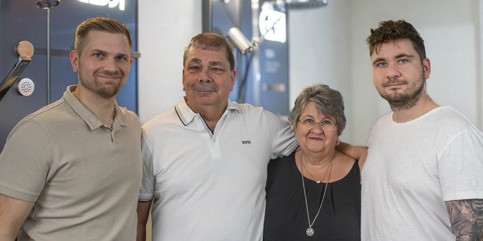 Eine Gruppe von vier Menschen, darunter zwei Männer und zwei Frauen, posiert freundlich vor einem stilvollen Hintergrund.