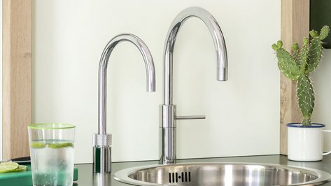 Ein moderner Wasserhahn mit zwei Ausläufen steht über einer glänzenden Spüle, umgeben von einer minimalistischen Kücheneinrichtung.