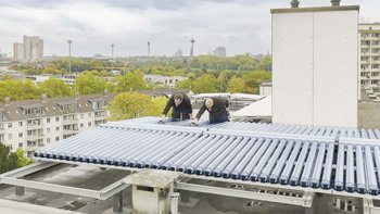 Zwei Arbeiter installieren Sonnenkollektoren auf einem Dach, während die Stadtlandschaft im Hintergrund sichtbar ist.