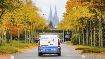 Ein Lieferwagen fährt eine von goldenen Herbstbäumen gesäumte Straße entlang, während die Kölner Domspitzen im Hintergrund sichtbar sind.