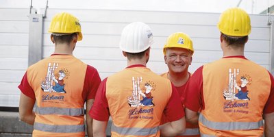Vier Handwerker stehen mit gelben Helmen und leuchtenden Westen in einer Baustellenszene, lächelnd und entspannt.