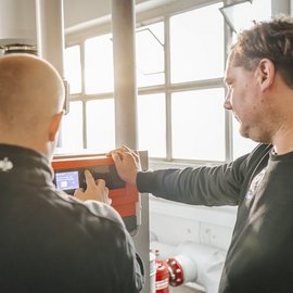 Zwei Männer stehen in einem hellen Raum und bedienen ein technisches Gerät, während sie konzentriert auf den Bildschirm schauen.