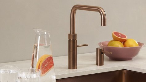 Ein eleganter Wasserhahn aus Kupfer steht neben einer Glaskaraffe mit Zitrusfrüchten und zwei Gläsern auf einem Tisch.