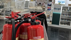 Eine Reihe roter Feuerlöscher steht in einem Einkaufswagen, während im Hintergrund eine Messe mit leeren Ständen sichtbar ist.