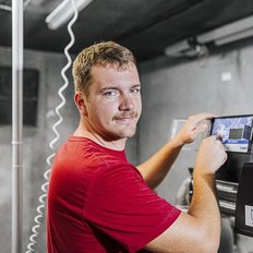 Ein junger Mann mit einem Schnurrbart trägt ein rotes T-Shirt und arbeitet an einer Maschine in einem modernen Raum.