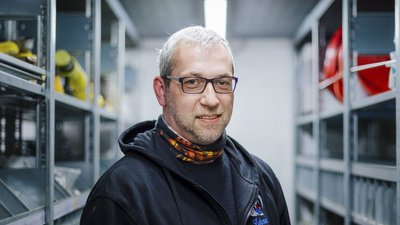 Ein Mann mit Brille und grauen Haaren lächelt in einem Lagerraum voller Regale mit Werkzeugen und Ausrüstung.