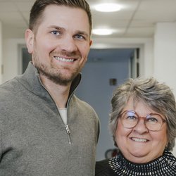 In einem modernen Flur posieren ein Mann mit blonden Haaren und eine Frau mit grauen Haaren, beide strahlen Freude und Zufriedenheit aus.