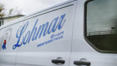 Ein weißer Lieferwagen mit dem Schriftzug 'Lohmar GmbH' und einem bunten Logo auf der Seite, vor einem unscharfen Hintergrund.