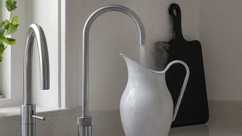 Ein eleganter Wasserkocher aus weißem Porzellan wird unter einem modernen Wasserhahn gefüllt, während Dampf aufsteigt.