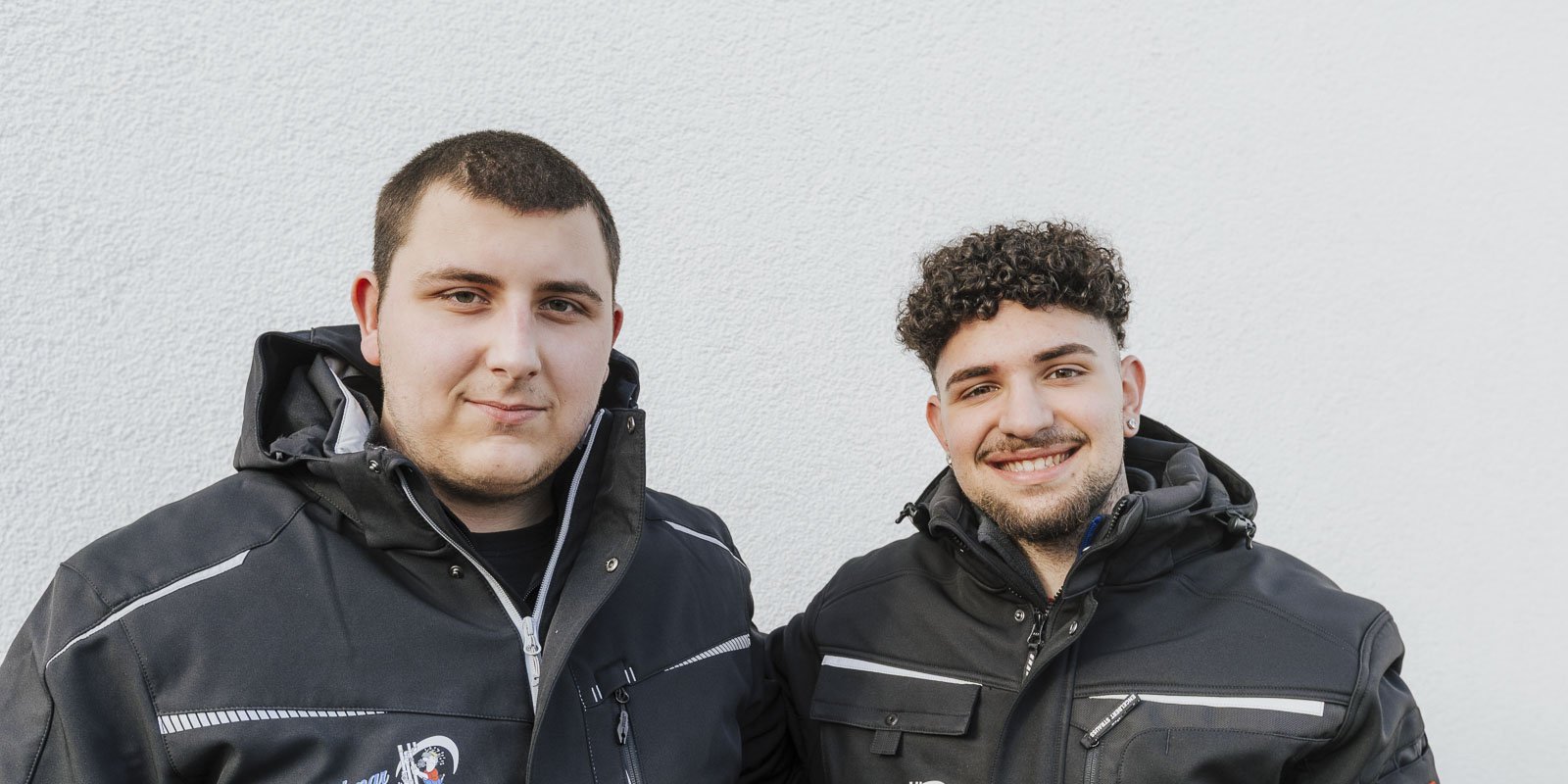 Zwei junge Männer stehen lächelnd vor einer hellen Wand, beide tragen schwarze Jacken mit Reißverschlüssen.