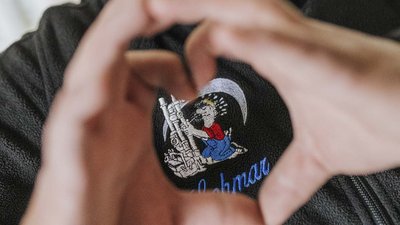 Zwei Hände formen ein Herz, während ein farbenfroher Aufnäher mit einem Cartoon-Charakter auf einem schwarzen Stoff sichtbar ist.