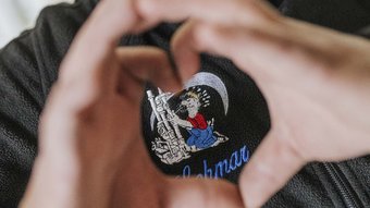 Herzformende Hände über einem kreativen Aufnäher Zwei Hände formen ein Herz, während ein farbenfroher Aufnäher mit einem Cartoon-Charakter auf einem schwarzen Stoff sichtbar ist.