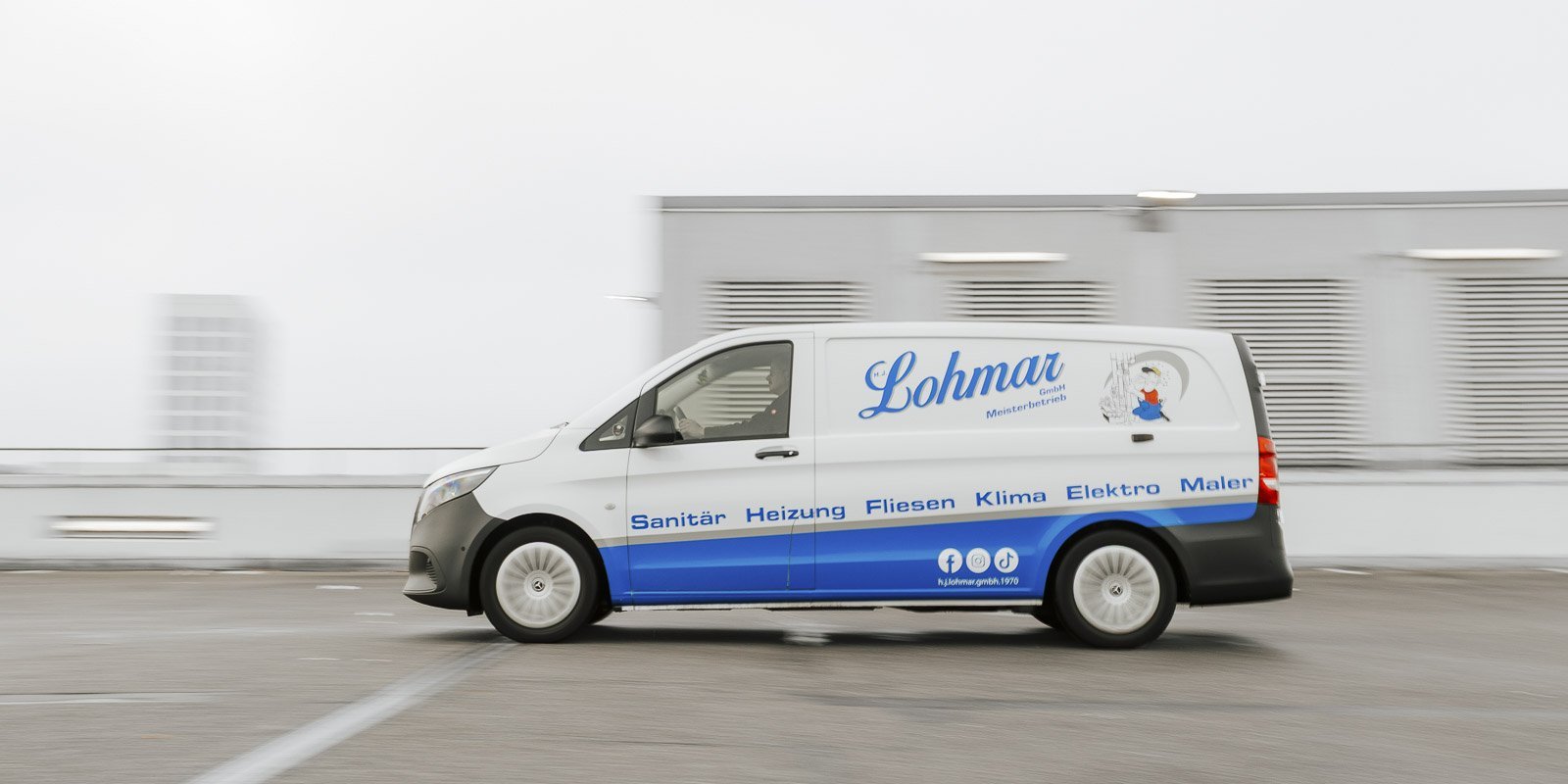 Ein Lieferwagen mit dem Logo von Lohmar fährt schnell über einen Parkplatz, umgeben von einer grauen, bewölkten Umgebung.