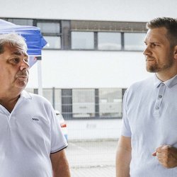 Zwei Männer stehen im Freien und unterhalten sich, während ein blauer Pavillon im Hintergrund sichtbar ist.
