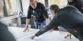 In einem hellen Raum analysieren Männer einen technischen Plan, ihre Gesten zeigen Engagement und Teamarbeit.