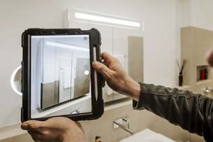 Eine Person hält ein Tablet, um einen modernen Badezimmerbereich mit eleganten Fliesen und Handtüchern zu fotografieren.
