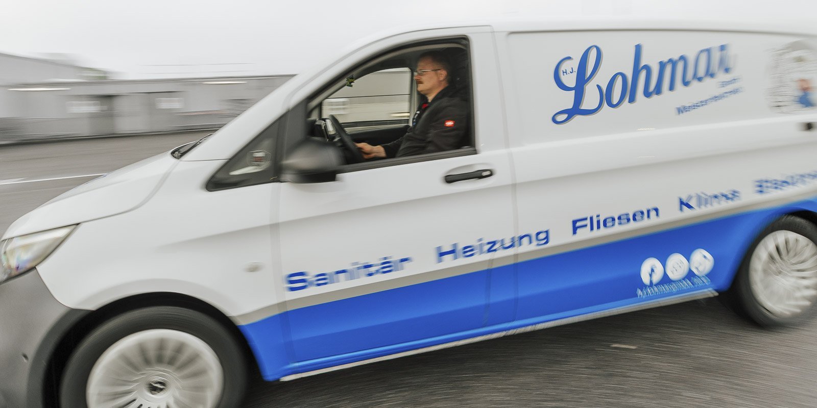 Der Fahrer eines Lieferwagens mit dem Logo 'Lohmar' ist unterwegs, während die Umgebung verschwommen ist.