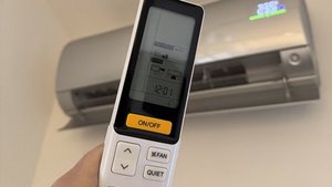 Eine Hand hält eine Fernbedienung für eine Klimaanlage, während die Klimaanlage an der Wand sichtbar ist.