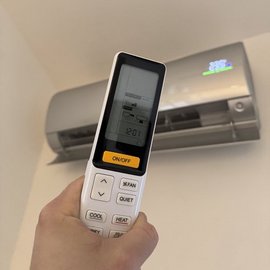 Eine Hand hält eine Fernbedienung für eine Klimaanlage, während die Klimaanlage an der Wand sichtbar ist.
