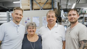 Vier Personen stehen lächelnd in einem Lagerraum, umgeben von Regalen und Materialien, die eine freundliche Atmosphäre schaffen.
