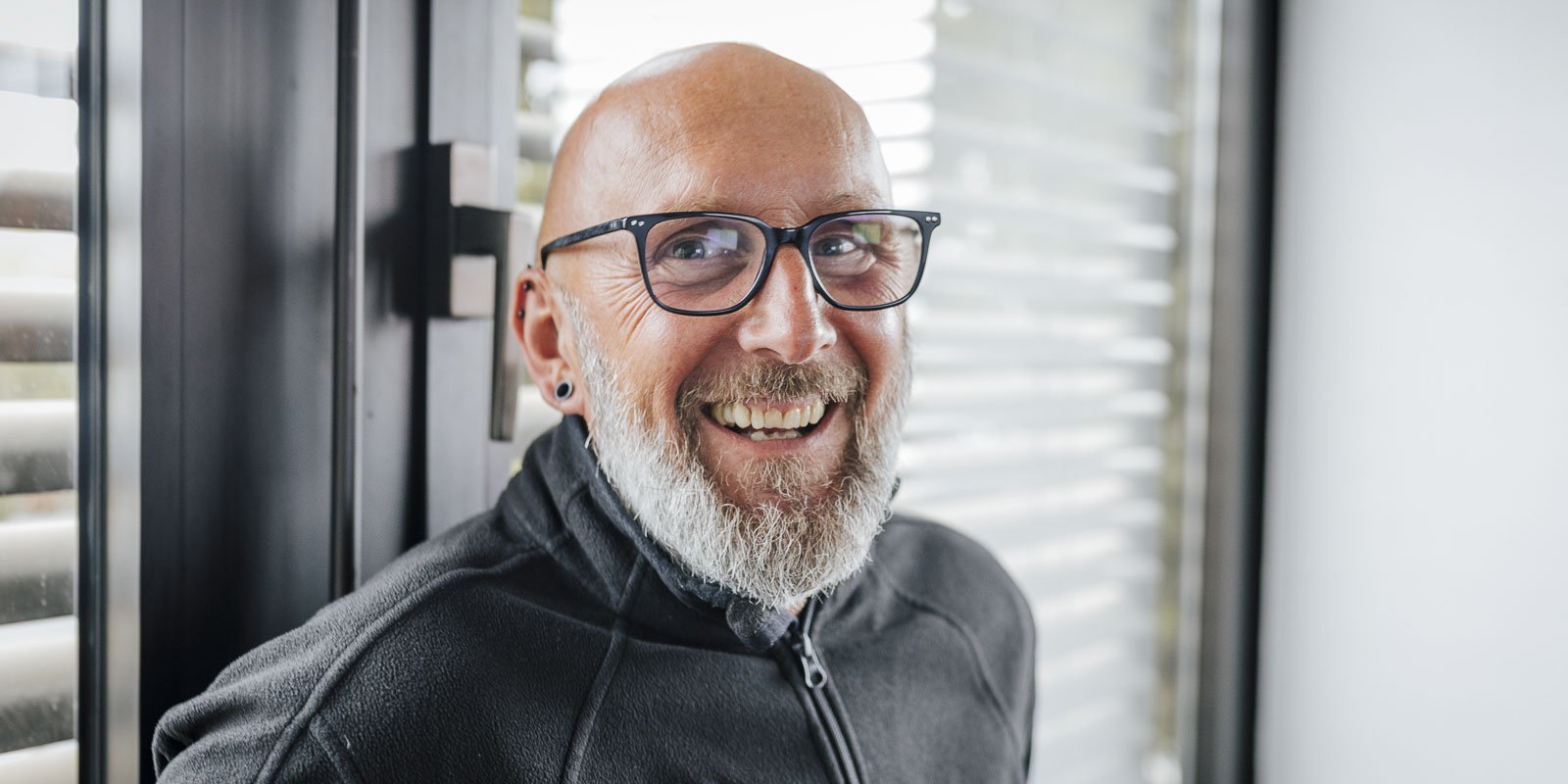 Ein lächelnder Mann mit Brille und Bart steht an einem Fenster mit Jalousien, strahlt Freude und Zufriedenheit aus.