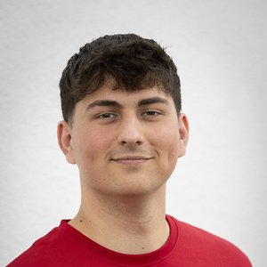 Ein junger Mann mit dunklen, lockigen Haaren lächelt selbstbewusst in die Kamera, während er ein rotes T-Shirt trägt.