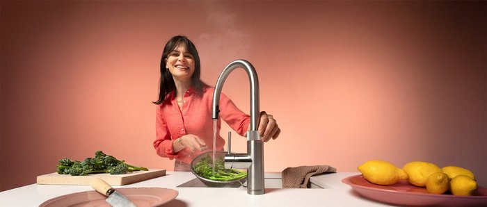 Eine lächelnde Frau in einem rosa Hemd wäscht frisches Gemüse unter einem modernen Wasserhahn in einer hellen Küche.