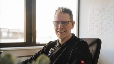 Ein Mann mit Brille lächelt freundlich in einem modernen Büro, während er an einem Schreibtisch sitzt und in die Kamera schaut.