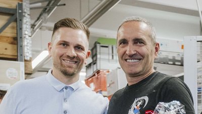Zwei Männer lächeln freundlich in einem modernen Raum, umgeben von Regalen und Arbeitsmaterialien.