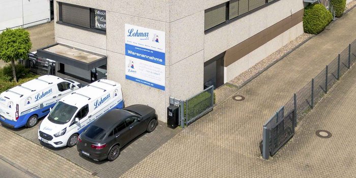 Zwei Lieferwagen mit dem Logo von Lohmar parken vor einem modernen Gebäude mit einem klaren blauen Schild zur Warenannahme.