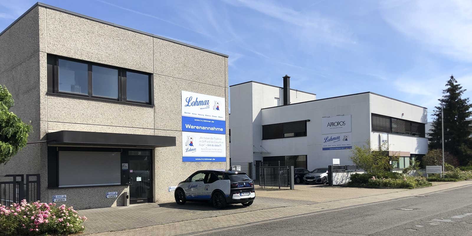 Zwei moderne Gebäude mit klaren Linien, eines mit einem großen Schild für Lohmar und das andere für Apropos.