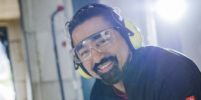 Ein lächelnder Mann mit Schutzbrille und Gehörschutz steht in einer hellen Werkstatt, bereit für die Arbeit.