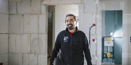 Ein lächelnder Mann steht in einem Baustellenbereich mit unvollendeten Wänden und sichtbaren elektrischen Installationen.