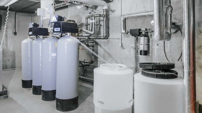 In einem modernen Technikraum stehen mehrere große, graue Wassertanks neben Rohren und einem schwarzen Filtergerät.