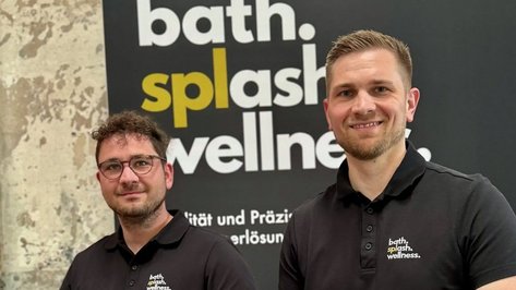 Zwei Männer in schwarzen Poloshirts lächeln vor einer Wand mit dem Schriftzug 'bath. splash. wellness.'