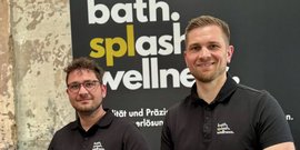 Zwei Männer in schwarzen Poloshirts lächeln vor einer Wand mit dem Schriftzug 'bath. splash. wellness.'
