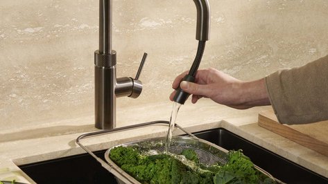 Eine Hand hält einen flexiblen Wasserhahn, während frisches Gemüse in einem Sieb gewaschen wird.
