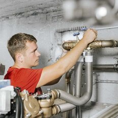 Ein Handwerker in einem roten Shirt arbeitet konzentriert an einer Rohrleitung in einem modernen Technikraum.