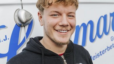 Ein junger Mann mit lockigem Haar lächelt freundlich vor einem weißen Lieferwagen mit blauer Schrift.