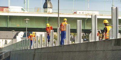 Mehrere Bauarbeiter arbeiten konzentriert an einem Geländer, während ein Fahrzeug über die Brücke fährt und die Sonne scheint.