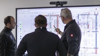 Drei Männer betrachten einen digitalen Bauplan auf einem großen Bildschirm, während einer mit einem Stift darauf zeigt.
