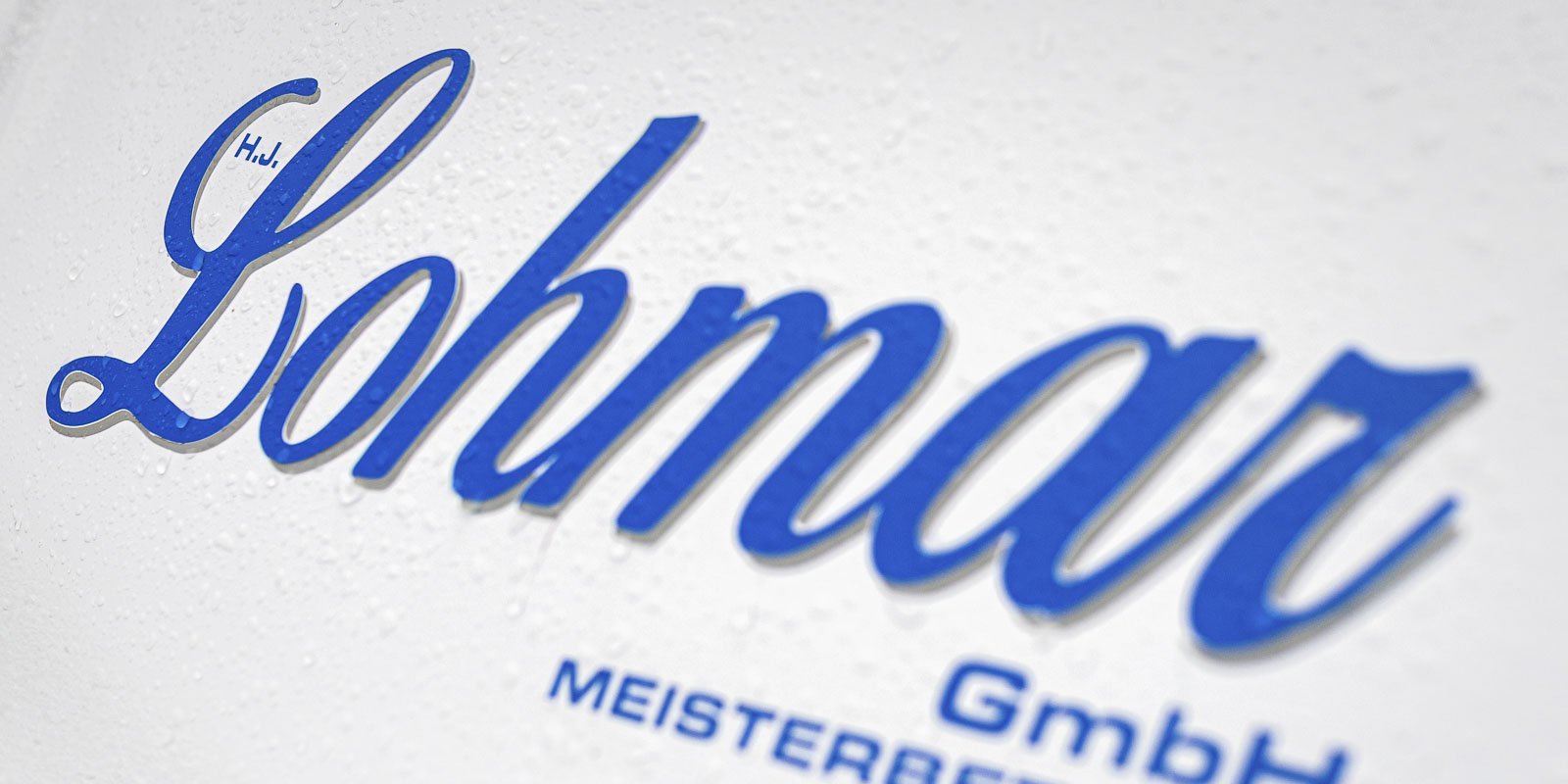 Das Logo von Lohmar GmbH in kräftigem Blau auf einer weißen, leicht feuchten Oberfläche, die die Schrift hervorhebt.