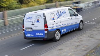 Lohmar Sanitärfahrzeug in Bewegung auf der Hauptstraße Ein Lieferwagen mit dem Logo von Lohmar fährt schnell auf einer Straße, umgeben von Bäumen und einem Zaun im Hintergrund.