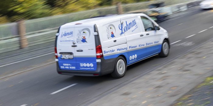 Ein Lieferwagen mit dem Logo von Lohmar fährt schnell auf einer Straße, umgeben von Bäumen und einem Zaun im Hintergrund.