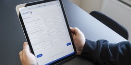 Das Bild zeigt eine Hand, die ein Tablet mit einem Online-Formular für Heizungsinformationen bedient, auf einem Tisch.