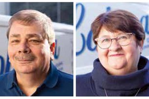 Die beiden Personen, ein Mann mit Schnurrbart und eine Frau mit Brille, zeigen ein herzliches Lächeln vor einem Lieferwagen.