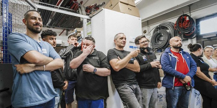 Eine Gruppe von Männern und Frauen steht in einem Raum, einige mit Getränken in der Hand, während sie aufmerksam zuhören.